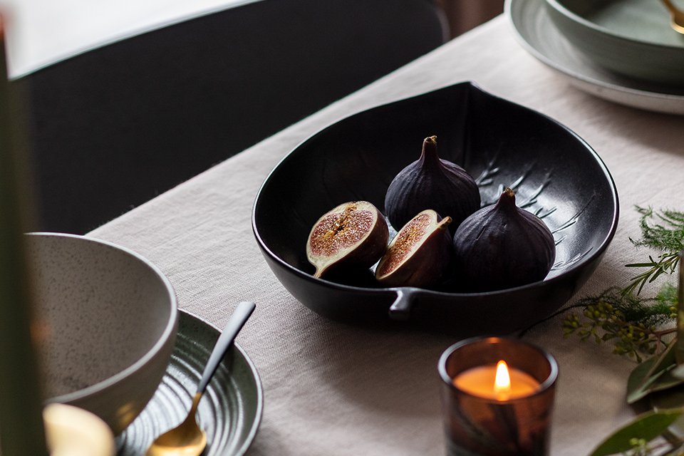 A black leaf shaped serving bowl on a dining table with figs in it.